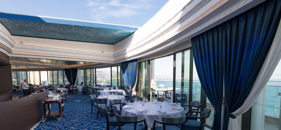 A bright and luxurious dining area with large windows offering expansive ocean views. The room features a blue and white color scheme, elegantly set tables, blue patterned carpet, dark blue curtains, and a ceiling with an artistic design. Sur mesure touches ensure some tables are delicately set for service.