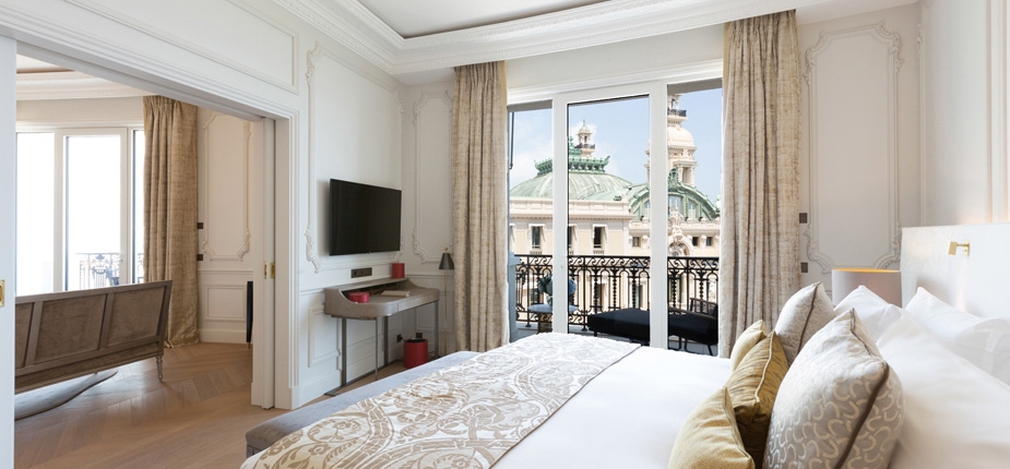 A luxurious hotel suite featuring an elegant bedroom with a large bed and decorative pillows. A wall-mounted TV, desk, and sofa are visible. Opening to a balcony with a view of a historic building with dome-shaped architecture, the room boasts sur mesure amenities for ultimate luxe. Light drapes frame the windows.