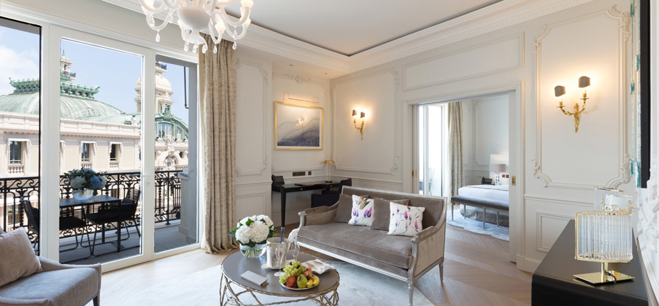 Elegant living room with white walls, wainscoting, and high ceilings, featuring a chandelier, sofa, and modern coffee table adorned with flowers and fruit. French doors open to a balcony with views of an ornate building. The boutique-style agencement extends to the bedroom visible through the doorway.