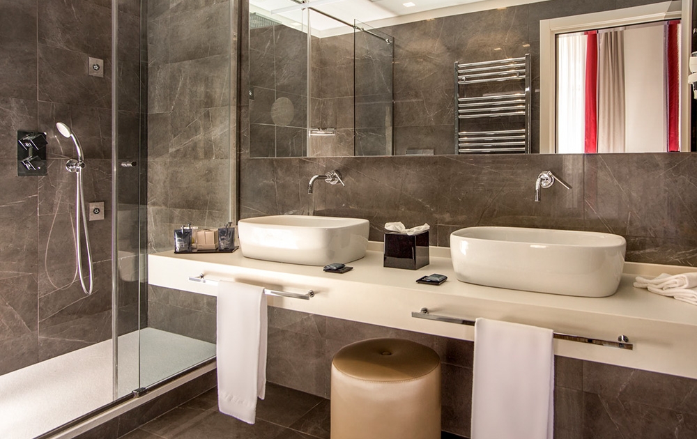 Salle de bains moderne avec carrelage marron, comprenant une douche à l'italienne en verre, deux vasques blanches sur un plan de travail beige, un grand miroir et un porte-serviettes chromé. Un tabouret rond beige et des serviettes blanches sont situés près des deux lavabos, le tout conçu avec une précision sur mesure pour une élégance de boutique.