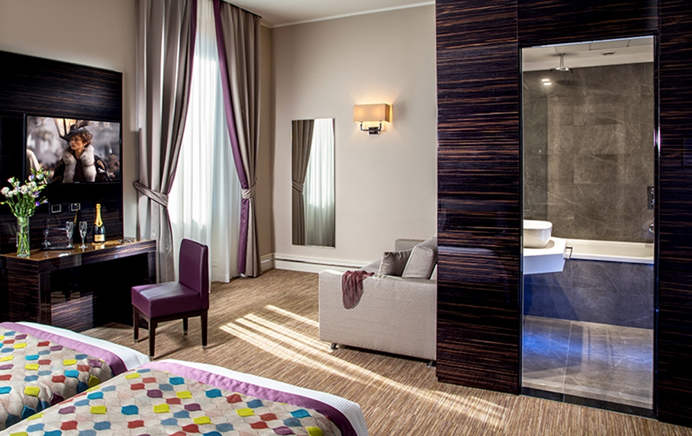 Chambre d'hôtel de charme avec deux lits jumeaux dotés de housses de couette colorées, un bureau et une chaise en bois foncé, une télévision à écran plat fixée au mur et un canapé. La chambre comprend également une grande fenêtre avec des rideaux, un miroir mural et une vue sur la salle de bains à travers une porte coulissante en verre.