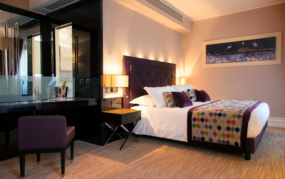 Une chambre d'hôtel-boutique moderne avec un grand lit doté d'une couverture à motifs colorés et d'oreillers décoratifs violets. L'ameublement de l'hôtel comprend un bureau et une chaise sombres, une table de nuit avec une lampe, des œuvres d'art murales et une vue partielle sur une salle de bains luxueuse avec un panneau en verre.