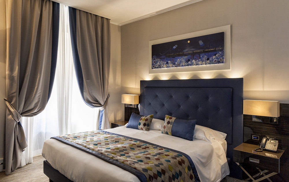 Une chambre d'hôtel-boutique moderne dotée d'un lit double soigneusement conçu avec une tête de lit bleu marine capitonnée et des oreillers et plaids colorés. Des rideaux doux aux tons neutres couvrent partiellement une grande fenêtre et des lampes élégantes ornent les tables de chevet. Une œuvre d'art représentant un paysage est suspendue au-dessus du lit, complétant l'agencement bien pensé.