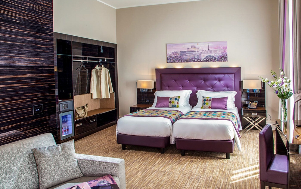 Une chambre d'hôtel-boutique moderne avec lits jumeaux, avec de luxueuses têtes de lit capitonnées violettes et des oreillers géométriques colorés. La chambre comprend un coin salon moelleux, une armoire, un bureau avec un vase de fleurs blanches et des œuvres d'art encadrées au-dessus des lits. Un éclairage chaleureux crée une atmosphère chaleureuse.