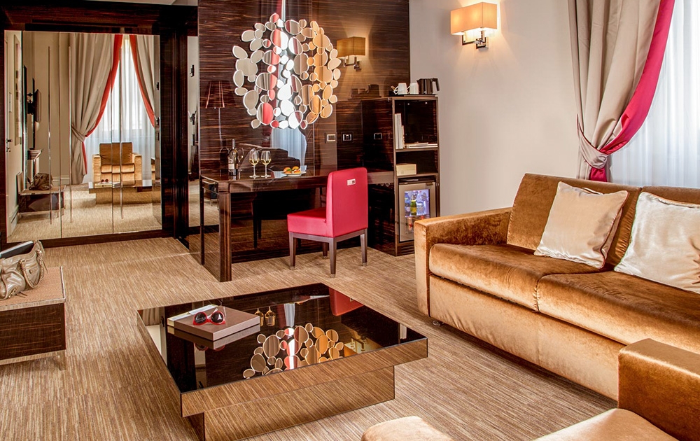 Chambre d'hôtel élégante avec un coin salon confortable. La chambre dispose d'un canapé beige luxueux avec des coussins blancs, d'une table basse en verre et d'un bureau avec une chaise rouge. Une œuvre d'art murale circulaire unique est suspendue au-dessus du bureau. La décoration de la chambre comprend un tapis beige et des rideaux aux accents rouges.