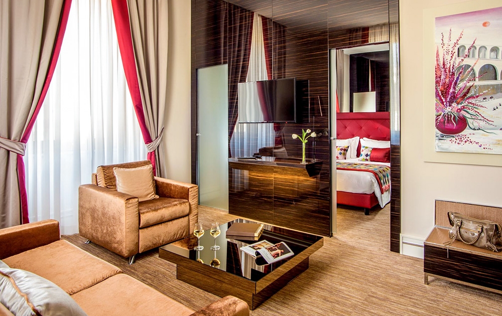 Suite d'hôtel moderne avec un coin salon avec des canapés beiges, une table basse en verre avec des magazines et des verres à vin, et une télévision à écran plat sur un meuble mural en bois. La chambre, visible à travers une élégante cloison vitrée d'Aménagement Hôtel Luxe, dispose d'un lit avec une tête de lit rouge et une literie colorée.