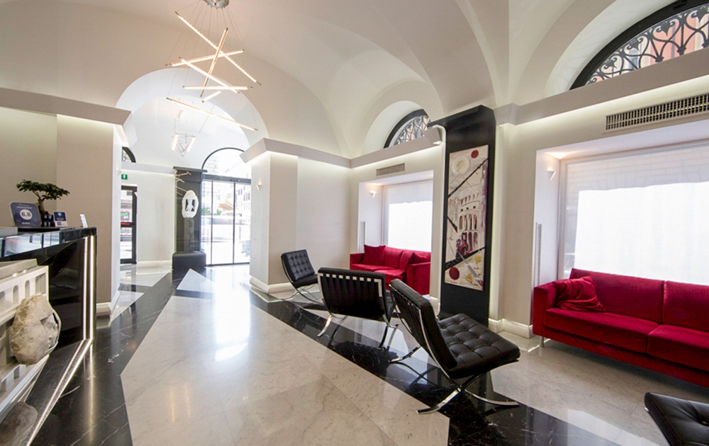 Un hall d'entrée moderne avec de hauts plafonds et des portes cintrées présente un sol noir et blanc, des canapés rouges, des chaises en cuir noir et un éclairage contemporain. L'espace boutique bénéficie de grandes fenêtres qui permettent à la lumière naturelle d'illuminer la zone, mettant en valeur son agencement sur mesure.