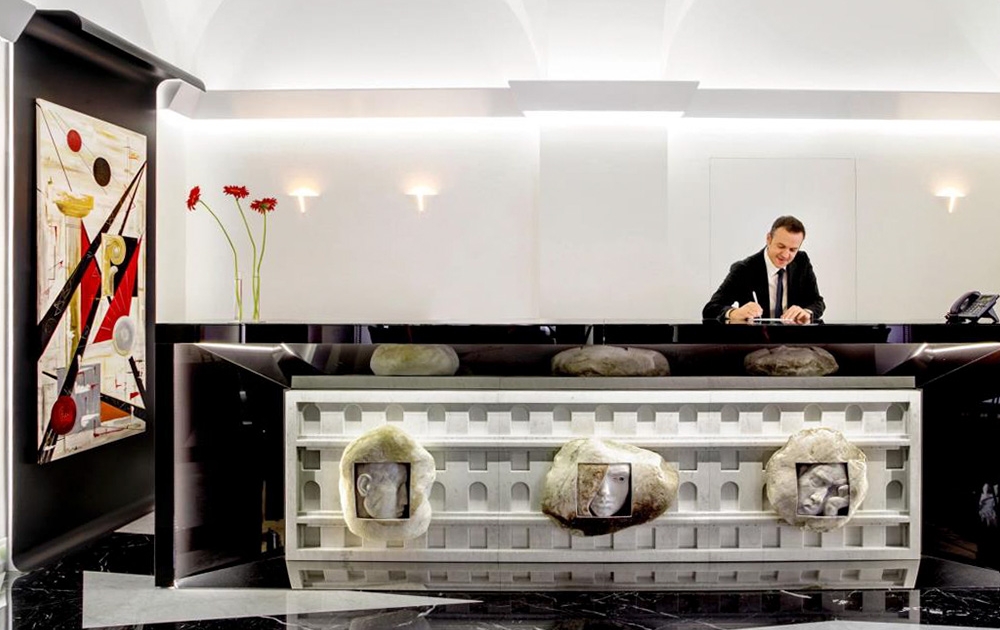 La réception d'un hôtel moderne est décorée d'une réceptionniste en costume noir qui travaille à un élégant bureau noir et blanc. Ce bureau sur mesure, qui met en valeur un savoir-faire artisanal sur mesure, est orné de sculptures artistiques en pierre. La toile de fond comprend un décor minimaliste avec des fleurs rouges et de l'art abstrait sur le mur.