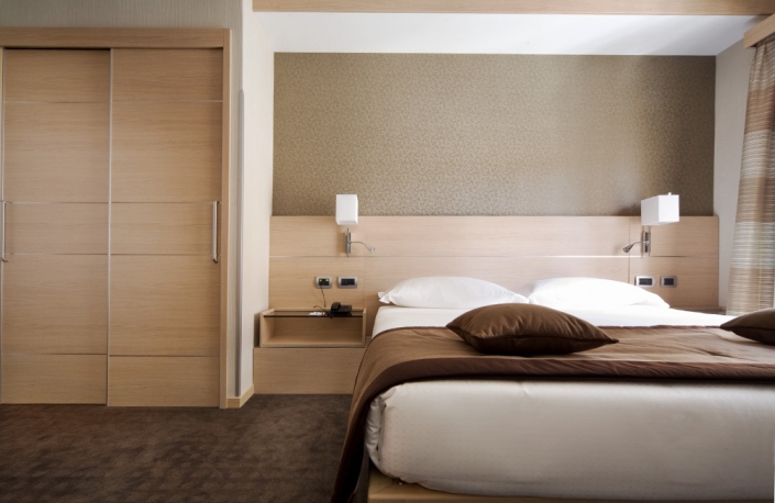 Una moderna camera da boutique hotel con un grande letto matrimoniale ricoperto di lino bianco e una coperta marrone, completata da due cuscini marroni. La camera è dotata di una testiera in legno, lampade da comodino, una piccola mensola con un telefono, un armadio con ante scorrevoli e una finestra ornata da tende trasparenti.