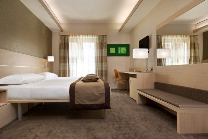 Una moderna camera d'albergo che mette in mostra un design sur mesure con un grande letto rivestito di biancheria da letto marrone e bianca, una TV a parete e una scrivania con una sedia arancione. Un'illuminazione accogliente esalta i colori neutri e grandi finestre con tende a fantasia lasciano entrare la luce naturale.