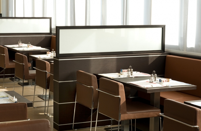 Un interno vuoto e moderno di un ristorante con panche e sedie in pelle marrone. I tavoli sono apparecchiati con posate, tovaglioli, saliere e pepiere e ciotole per condimenti. Una luce soffusa filtra attraverso le tende trasparenti che coprono le finestre sullo sfondo, mettendo in mostra un design di ristorante di hotel di lusso su misura.