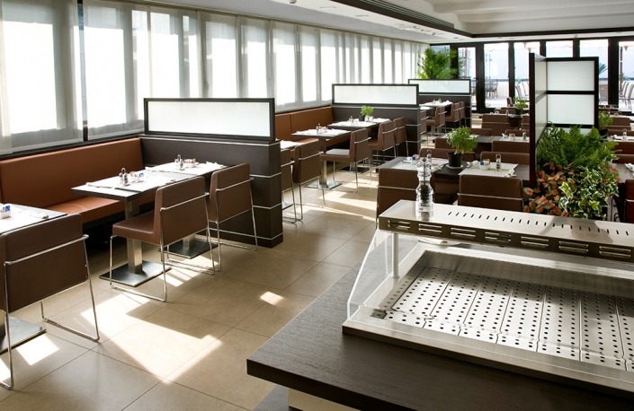 Un ristorante moderno e soleggiato con sedie in pelle marrone e tavoli disposti ordinatamente. Le sedute a panca fiancheggiano le finestre e ci sono piante che aggiungono un tocco di verde. Lo spazio è pulito e pronto per gli ospiti, con utensili sistemati su ogni tavolo e una grande griglia sur mesure in primo piano.