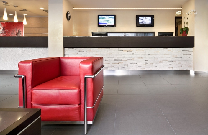 Moderna hall di un hotel con una poltrona in pelle rossa in primo piano. Il banco della reception, in legno scuro con facciata in mattoni bianchi, è dietro la sedia. Due televisori a schermo piatto e un orologio sono appesi alla parete sopra la scrivania. Questo spazio boutique è ben illuminato ed elegante, e offre un'esperienza su misura.
