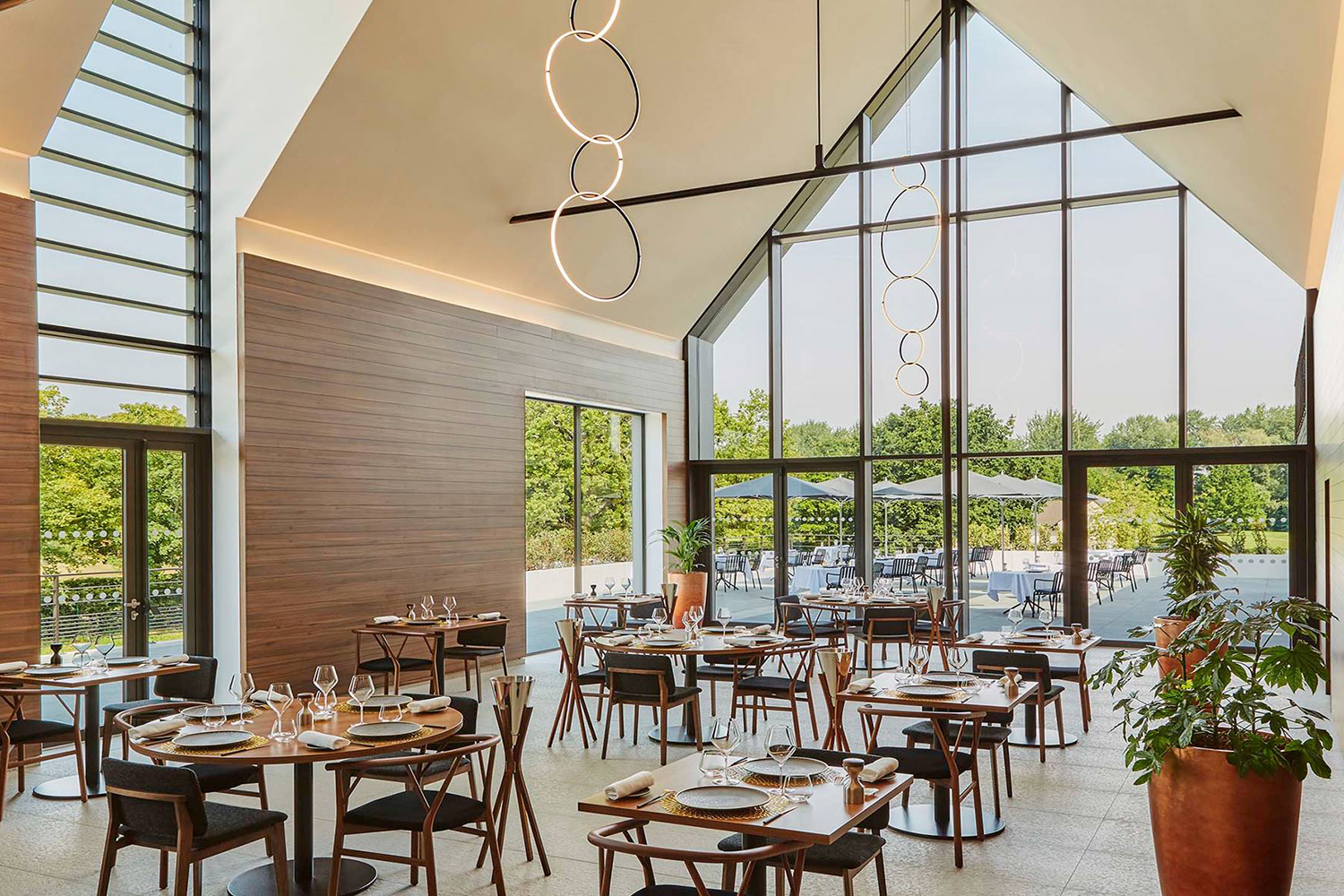 A modern, upscale restaurant with high vaulted ceilings, large floor-to-ceiling windows, and contemporary circular pendant lights. Wooden tables and chairs are neatly arranged, set for dining, with an outdoor patio visible through the glass doors—perfectly reflecting our sur mesure Amenagement hotel restaurant concept.
