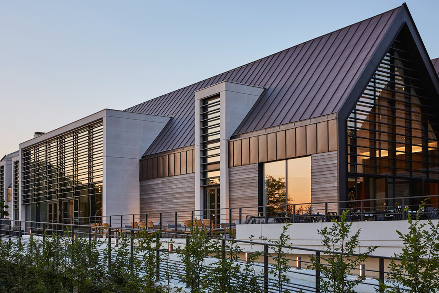 A modern building with a mix of concrete, glass, and metallic materials stands against a twilight sky. The structure features large windows, louvered panels, and a slanted roof designed with sur mesure precision. A metal railing runs along the side, with small trees planted in the foreground.