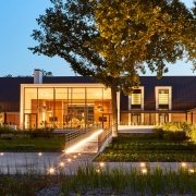 Un bâtiment moderne avec de grandes baies vitrées est illuminé au crépuscule, niché parmi de grands arbres et une végétation luxuriante. Un chemin, orné de petites lumières, mène à l'entrée. La conception architecturale présente des lignes épurées, du bois naturel et un toit sombre mettant en valeur un agencement de luxe.