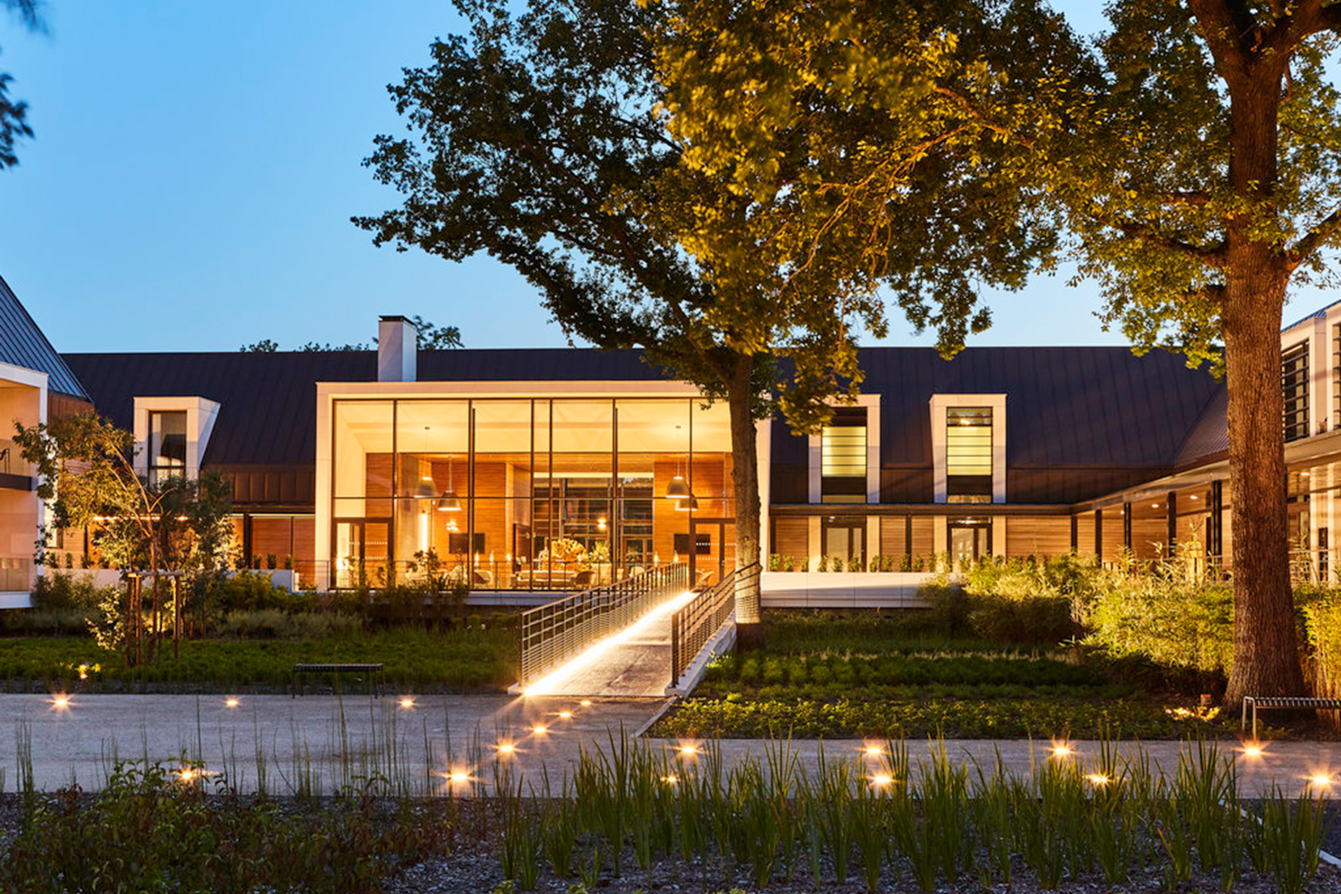 A modern building with large glass windows, illuminated at dusk, exudes a luxe charm. The entrance features a pathway flanked by small lights, reminiscent of a boutique hotel. Various plants and trees enhance the serene atmosphere, while the clear sky provides a peaceful backdrop.