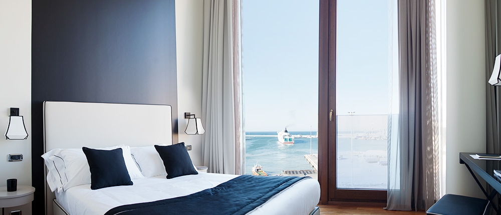 Une chambre moderne et luxueuse avec une grande fenêtre offrant une vue sur l'océan. La chambre comprend un lit double avec une literie noire et blanche, des murs blancs accentués par un mur noir, du parquet et des rideaux élégants encadrant la grande fenêtre. Un bureau et une chaise de style boutique sont visibles à droite.