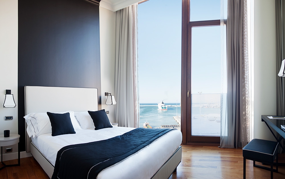 Modern hotel room with a large bed, black and white bedding, and two matching pillows. Floor-to-ceiling windows with light curtains reveal a stunning ocean view. The room features wooden flooring, a nightstand, and a sleek desk. The walls are a blend of black and white sur mesure agencement.
