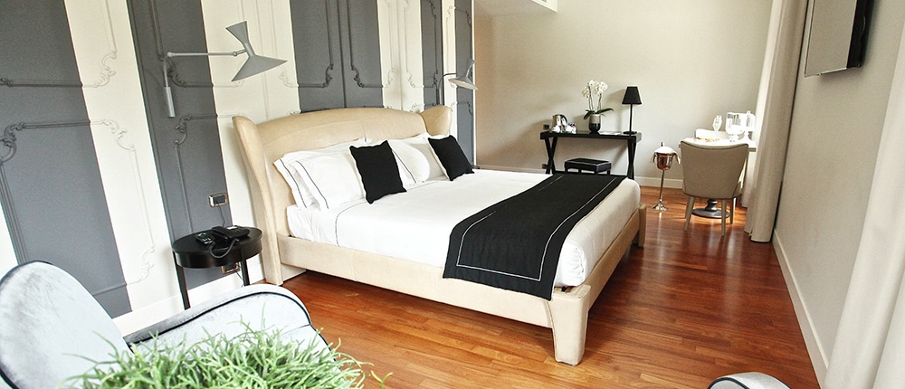 Une chambre d'hôtel moderne dispose d'un lit moelleux avec des draps noirs et blancs, entouré de meubles aux tons neutres et d'un parquet en bois. La chambre comprend un bureau, une chaise et des plantes décoratives. De grandes fenêtres permettent à la lumière naturelle de remplir l'espace, mettant en valeur le décor élégant et minimaliste et l'agencement de luxe sur mesure.