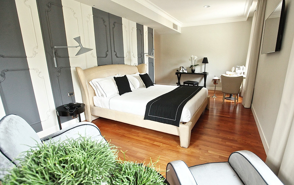 A modern hotel room with wooden flooring features a large bed with white linens and black accents, a desk with a chair, and a flat-screen TV mounted on the wall. There are two light gray armchairs and a small plant in the corner. The walls, inspired by luxe design from their contract division, boast an elegant pattern.