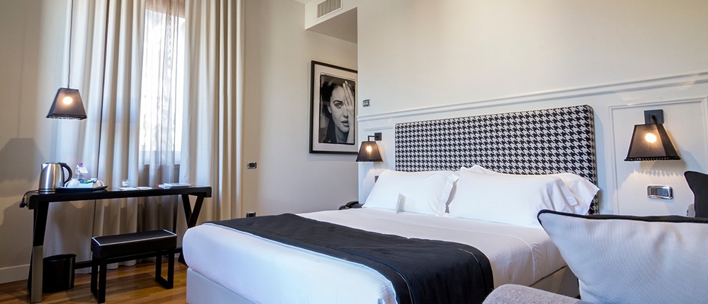 Une chambre d'hôtel moderne respire le luxe avec un grand lit doté d'une tête de lit à motif pied-de-poule et d'une literie noire et blanche. Elle comprend un bureau, une bouilloire, des tasses et une photo encadrée. Des rideaux transparents couvrent la fenêtre et des planchers en bois rehaussent l'éclairage ambiant chaleureux de cet espace hôtelier Aménagement haut de gamme.