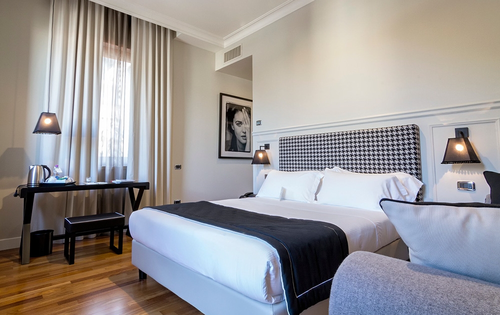 A modern hotel room with a large bed that has white linens and a black-and-white patterned headboard. There's a desk with amenities like a kettle, mugs, and a lamp to the left of the bed. A black-and-white portrait hangs above the desk. The room features light-colored curtains, showcasing thoughtful agencement.
