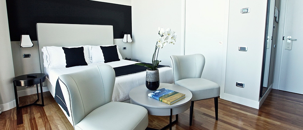 Une chambre d'hôtel moderne dotée d'un grand lit avec une tête de lit grise et des draps blancs, accentués par des oreillers noirs et un chemin de lit noir. Il y a deux fauteuils blancs et une table ronde avec un livre et une plante en pot, incarnant le design luxueux de l'hôtel Aménagement. La chambre a du parquet.