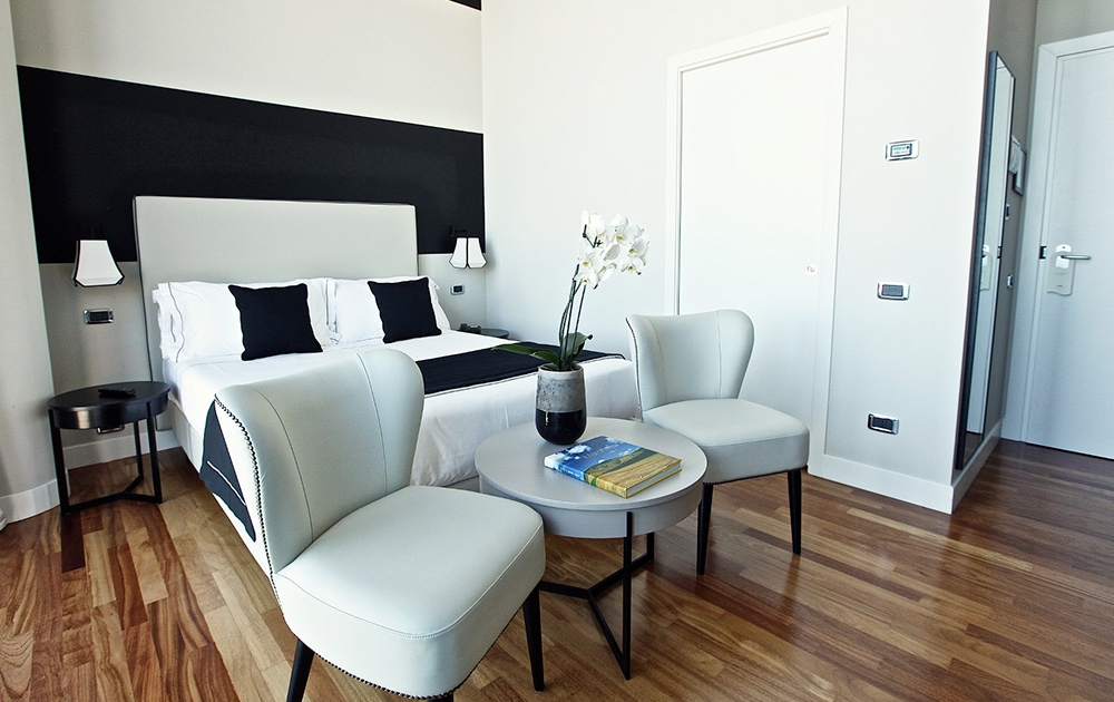 A modern, luxe hotel room with a white and black color scheme, featuring a double bed with black cushions, two white armchairs around a circular table, and a small bedside table with a lamp. The room has wooden flooring and a potted plant on the table.