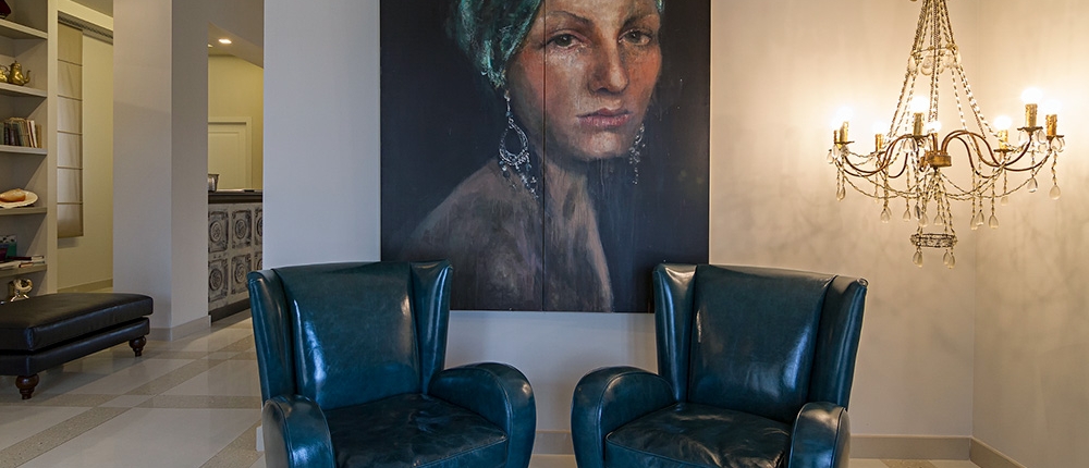 Une pièce moderne conçue par un expert en agencement présente deux fauteuils en cuir bleu canard sous le portrait d'une femme portant un foulard et des boucles d'oreilles. À droite, un lustre en cristal avec des ampoules allumées ajoute une touche de luxe à l'espace. Une porte ouverte et des étagères sont visibles en arrière-plan.
