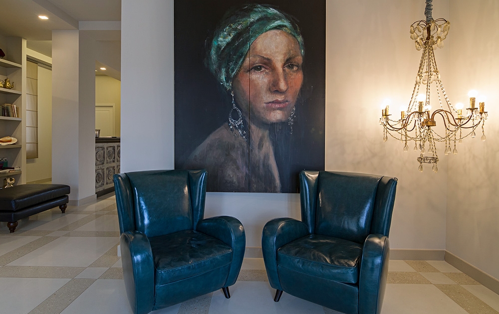A stylish room features two teal leather chairs facing a wall with a large, detailed portrait of a woman wearing a green headscarf. The painting is illuminated by a vintage chandelier on the right. Shelving and a black bench in the adjacent room add to the boutique ambiance.