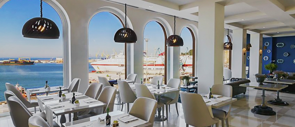 Un restaurant moderne et luxueux avec des tables blanches et des chaises grises est installé à l'intérieur près de grandes fenêtres cintrées. Les fenêtres offrent une vue sur un quai avec des bateaux et une mer bleue au-delà. Des suspensions noires sont suspendues au plafond et la pièce est bien éclairée par la lumière naturelle.