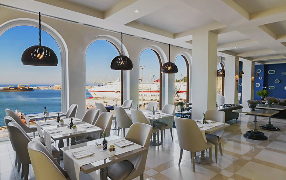 A stylish restaurant interior featuring light gray chairs, white tables, and black pendant lighting. Large arched windows provide a scenic view of a bustling harbor with ships docked under a clear blue sky. The modern agencement creates an inviting atmosphere reminiscent of a boutique hotel restaurant.