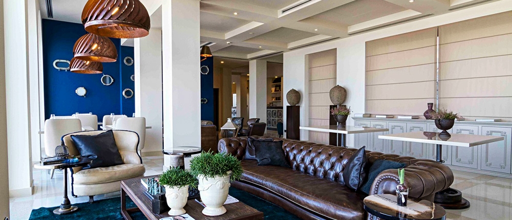 Un salon moderne avec un canapé en cuir marron, une table basse en verre ornée de plantes et des suspensions contemporaines dégage une atmosphère luxueuse. En arrière-plan, une salle à manger au design élégant avec un mur d'accent bleu et de gracieuses chaises blanches met en valeur l'aménagement expert de l'hôtel. L'espace est bien éclairé par la lumière naturelle.