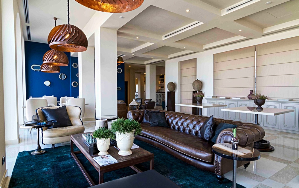 A modern living room features a brown leather sofa, green plants, and a dark blue rug. Elegant copper pendant lights hang above. In the background, a white dining area with a blue accent wall and framed art can be seen, along with various decorative items, giving it a boutique feel akin to high-end amenagement hotel restaurant interiors.