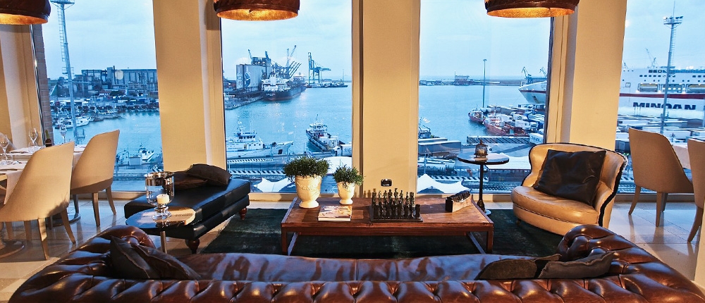 Un coin salon confortable, doté d'un canapé en cuir marron, de fauteuils et d'une table basse avec un jeu d'échecs, donne sur un port animé à travers trois grandes fenêtres cintrées. La décoration nautique et l'éclairage chaleureux créent une atmosphère relaxante, parfaite pour mettre en valeur un design réfléchi dans les espaces hôteliers.