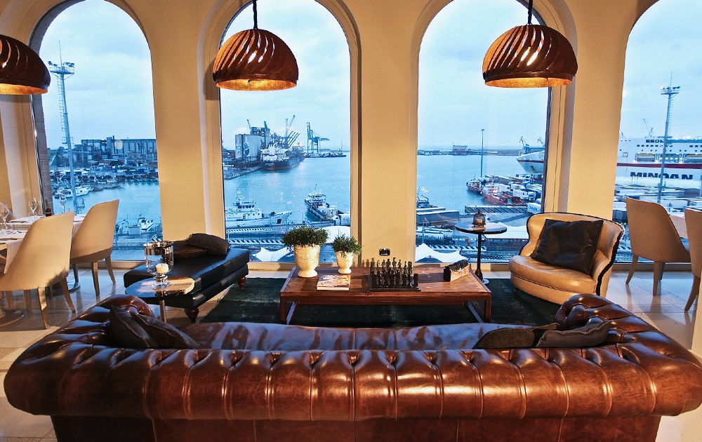 A luxurious living room with a brown leather sofa, a bespoke chess set on a wooden coffee table, and large arched windows providing a panoramic view of a busy waterfront and docks. Ships and cranes are visible outside. The room is warmly lit with pendant lights, embodying boutique elegance.