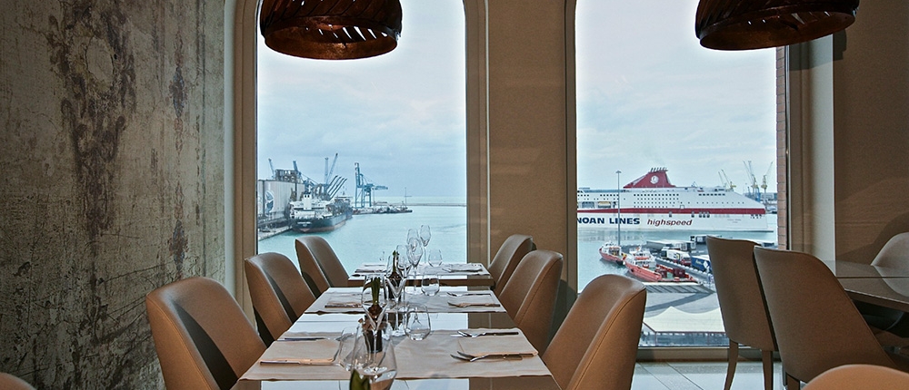 Un intérieur de restaurant bien éclairé avec une décoration moderne, avec des tables dressées avec des nappes blanches et de la verrerie. De grandes fenêtres cintrées offrent une vue panoramique sur un port, mettant en valeur des navires et des grues amarrés sur un ciel nuageux. L'ambiance luxueuse est renforcée par un agencement méticuleux dans tout l'espace.