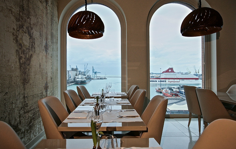 A modern restaurant interior featuring tables set for dining. Large arched windows provide a view of a bustling harbor with cranes and a large ferry docked. Pendant lights with a woven design hang from the ceiling, and natural light fills the space, showcasing expert agencement by our contract division.