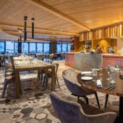Agencement hôtel, méticuleusement conçu par GH Partners, présente un plafond chaleureux en bois et un sol carrelé à motifs. Les tables sont dressées avec des verres et des assiettes, entourées de chaises moelleuses. Des étagères avec divers objets et un comptoir sont visibles en arrière-plan. De grandes fenêtres donnent sur un paysage enneigé.