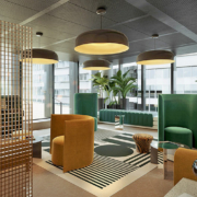Un salon de bureau moderne avec des fauteuils verts et marron, un tapis rayé et un petit tabouret à motif zébré. De grands plafonniers circulaires sont suspendus au-dessus, tandis que des plantes en pot près des fenêtres rehaussent la vue sur la ville. Une cloison en treillis se trouve à gauche, reflétant un « agencement sur mesure » : optimisez votre budget avec un partenaire unique.