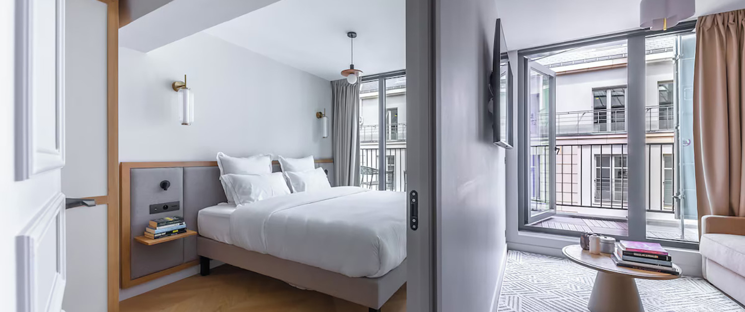 Chambre moderne à la Tour Pleyel – H4 Hotel Wyndham, avec un grand lit et une table de chevet, une literie blanche et des murs gris clair. Séjour ouvert avec portes-fenêtres donnant sur un balcon, canapé, table basse ronde et tapis à motifs gris sous un éclairage tamisé.