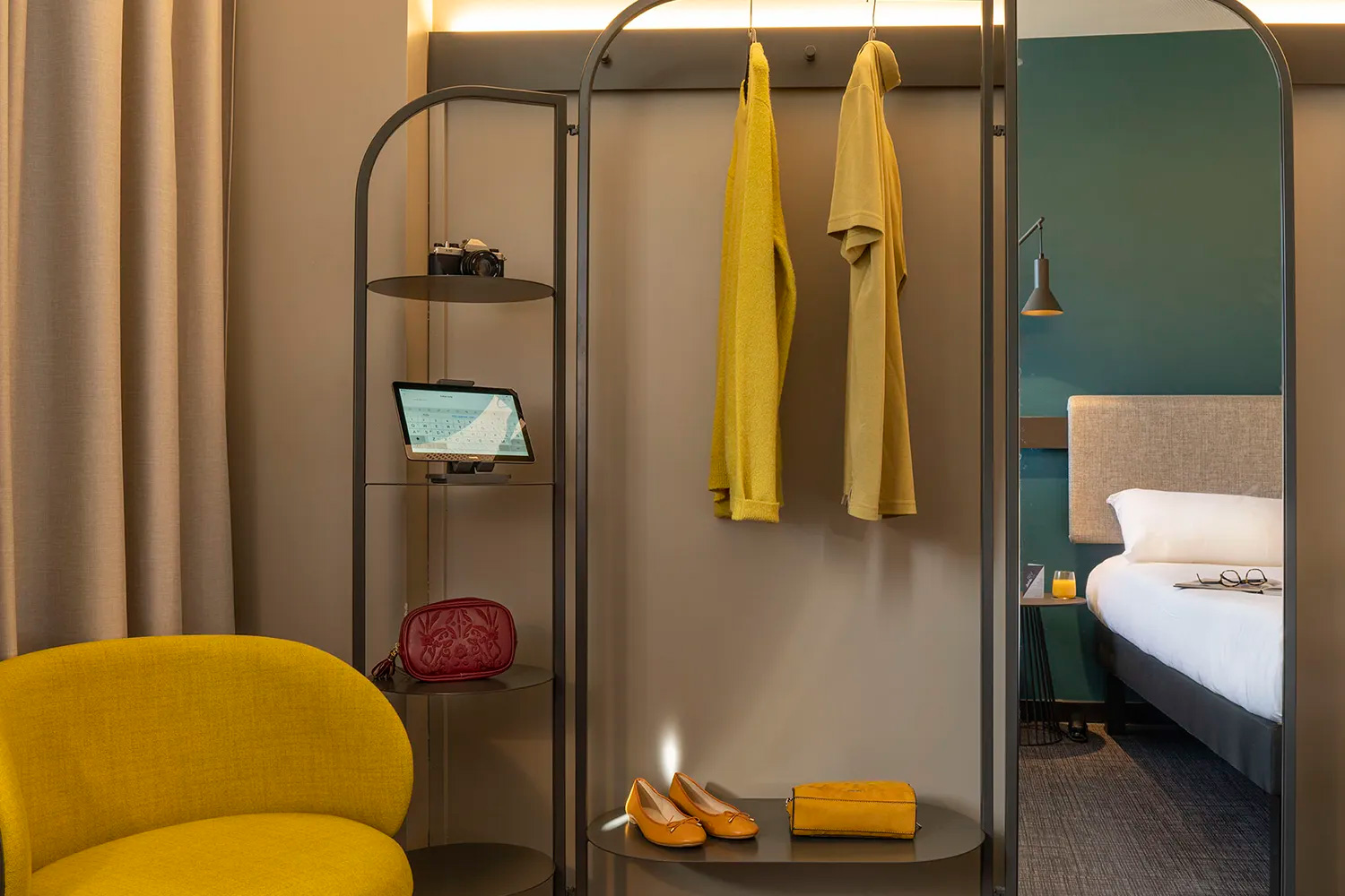 Une chambre d'hôtel moderne dispose d'un agencement sur mesure avec une chaise jaune, une armoire ouverte contenant des vêtements jaunes, des chaussures et un sac à main, des étagères affichant un sac rouge, un appareil photo et une tablette, et un lit partiellement visible en arrière-plan.