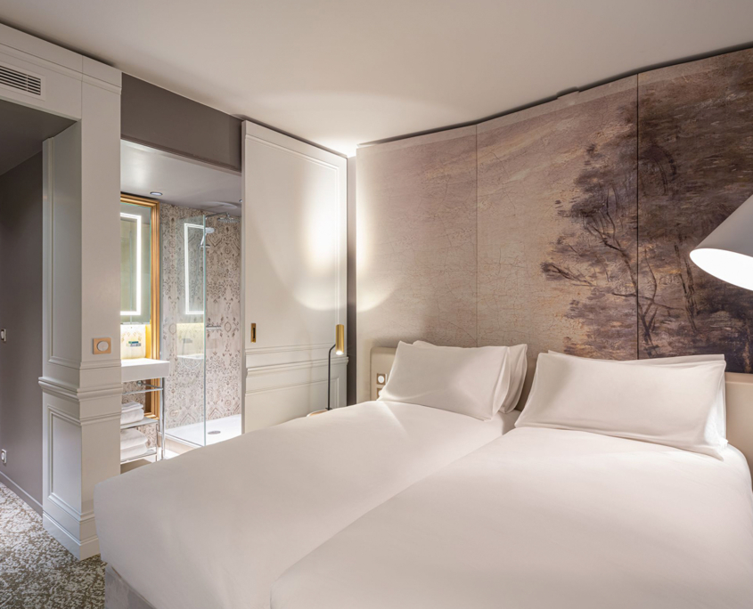 Una moderna camera d'albergo con due letti singoli ben fatti, un murale raffigurante un albero, un comodino con una lampada e un bagno con pareti in vetro e decorazioni bianche e beige visibili attraverso una porta scorrevole.