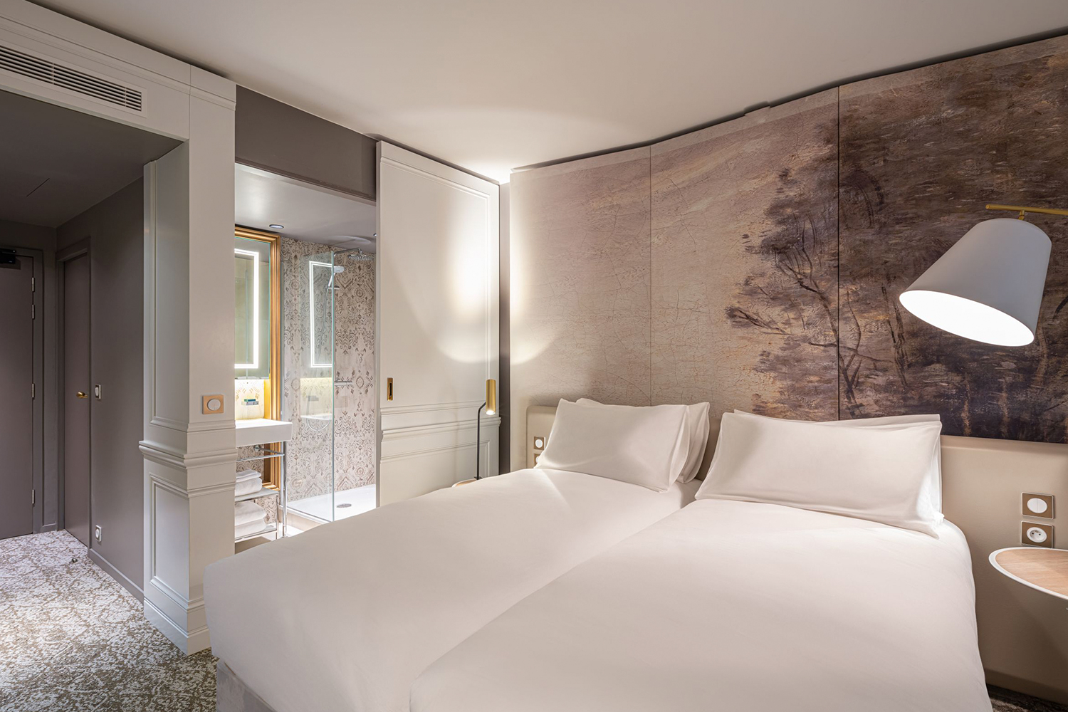 Una moderna camera d'albergo con due letti singoli ben fatti, un murale raffigurante un albero, un comodino con una lampada e un bagno con pareti in vetro e decorazioni bianche e beige visibili attraverso una porta scorrevole.