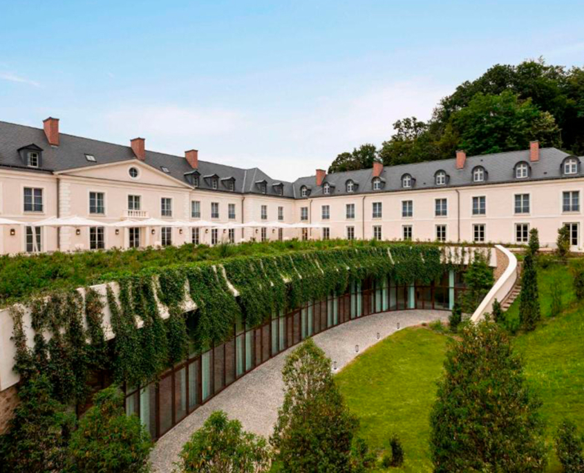 Un grande ed elegante edificio color crema, con tetto in ardesia e numerose finestre, si affaccia su una terrazza curva ricoperta di edera, circondata da prati verdi e alberi sotto un cielo azzurro e terso.