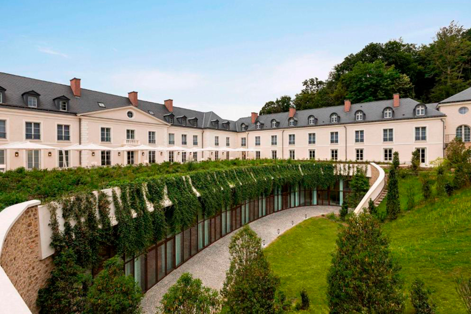 Un grande ed elegante edificio color crema, con tetto in ardesia e numerose finestre, si affaccia su una terrazza curva ricoperta di edera, circondata da prati verdi e alberi sotto un cielo azzurro e terso.