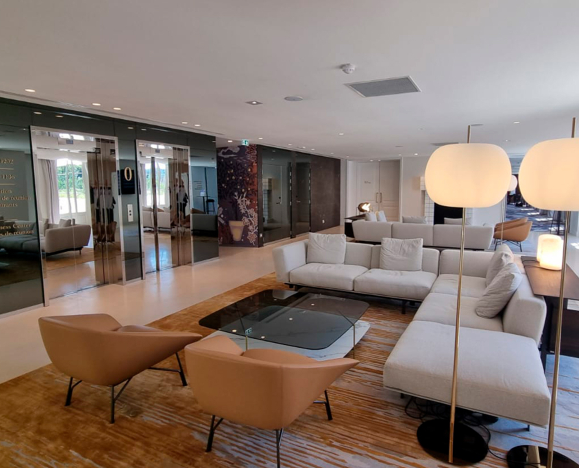 Moderna area lounge con poltrone beige, un grande divano componibile grigio chiaro, un tavolino da caffè in vetro, lampade da terra e pareti a specchio, impreziosita da un tappeto dai toni caldi e da abbondante luce naturale proveniente dalle finestre.