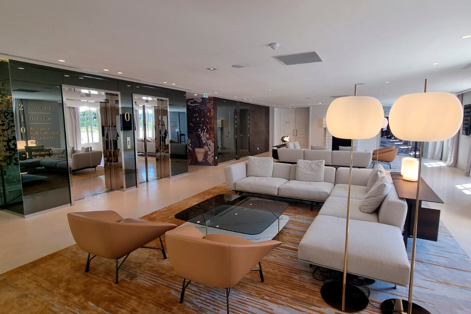 Moderna area lounge con poltrone beige, un grande divano componibile grigio chiaro, un tavolino da caffè in vetro, lampade da terra e pareti a specchio, impreziosita da un tappeto dai toni caldi e da abbondante luce naturale proveniente dalle finestre.