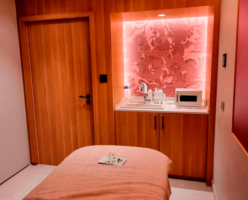 Un'accogliente sala per trattamenti spa con un lettino da massaggio coperto da un lenzuolo rosa, una porta e degli armadietti in legno, un bancone con prodotti da spa, un asciugamano e un microonde, il tutto illuminato da una calda luce rosa.